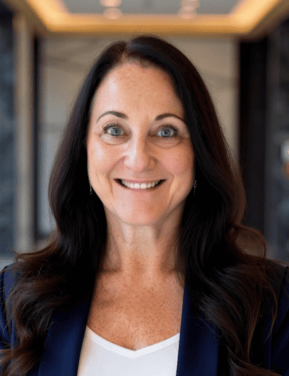 A forward-facing headshot of a smiling white woman with dark hair, wearing a black blazer and white shirt.