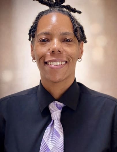 Direct, head-on photo of Commissioner Tanisha Arena, a Black Woman with dreads tied up, who is wearing a purple tie and black collared shirt.