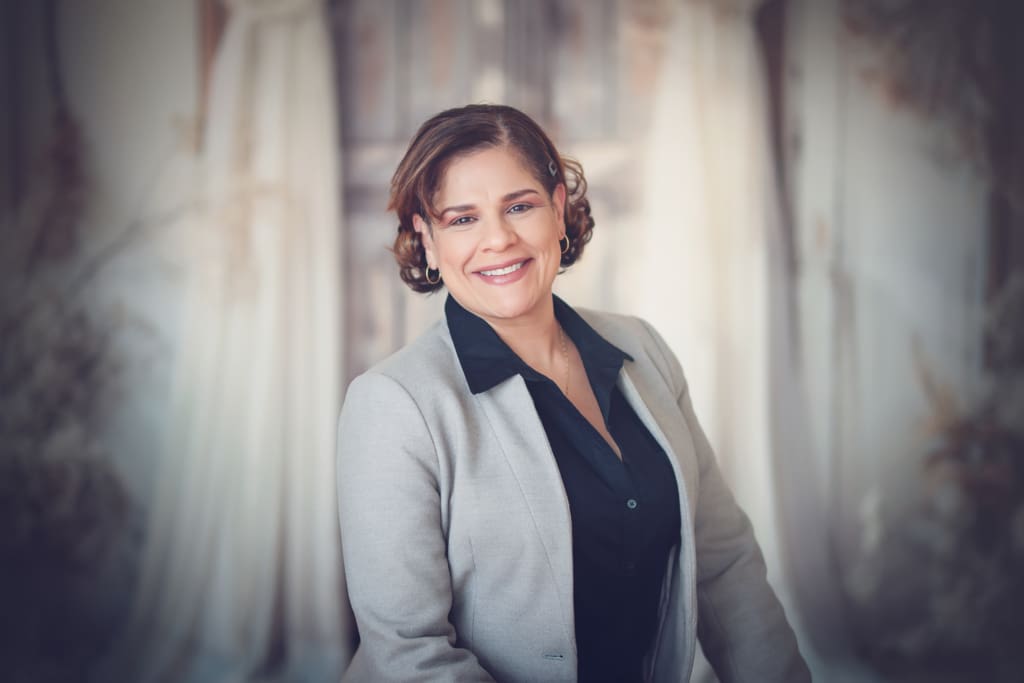 A smiling headshot of a middle-aged latina woman wearing a grey blazer and black collared shirt.