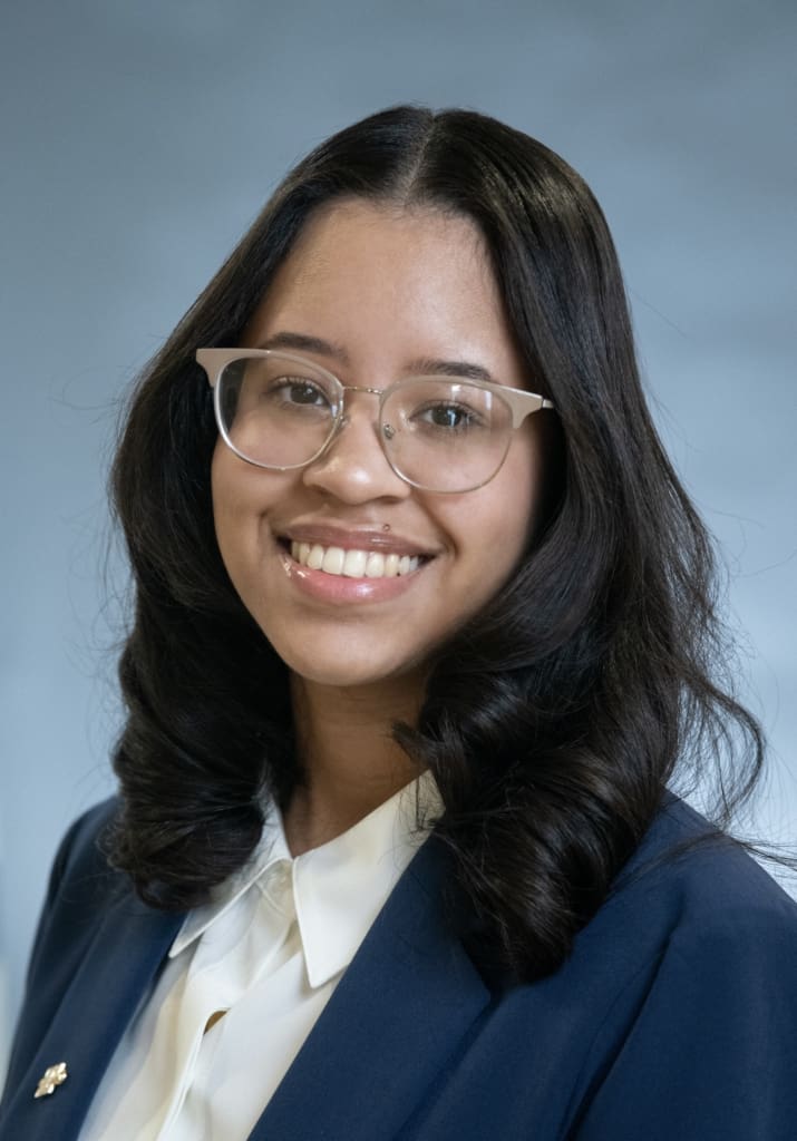 A smiling headshot of a young latina woman with glasses, wearing a blazer and white collared shirt.