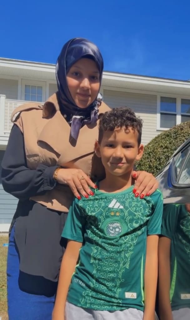 A smiling photo of a young woman wearing a hijab, posing with her hands on the shoulders of her young son. 