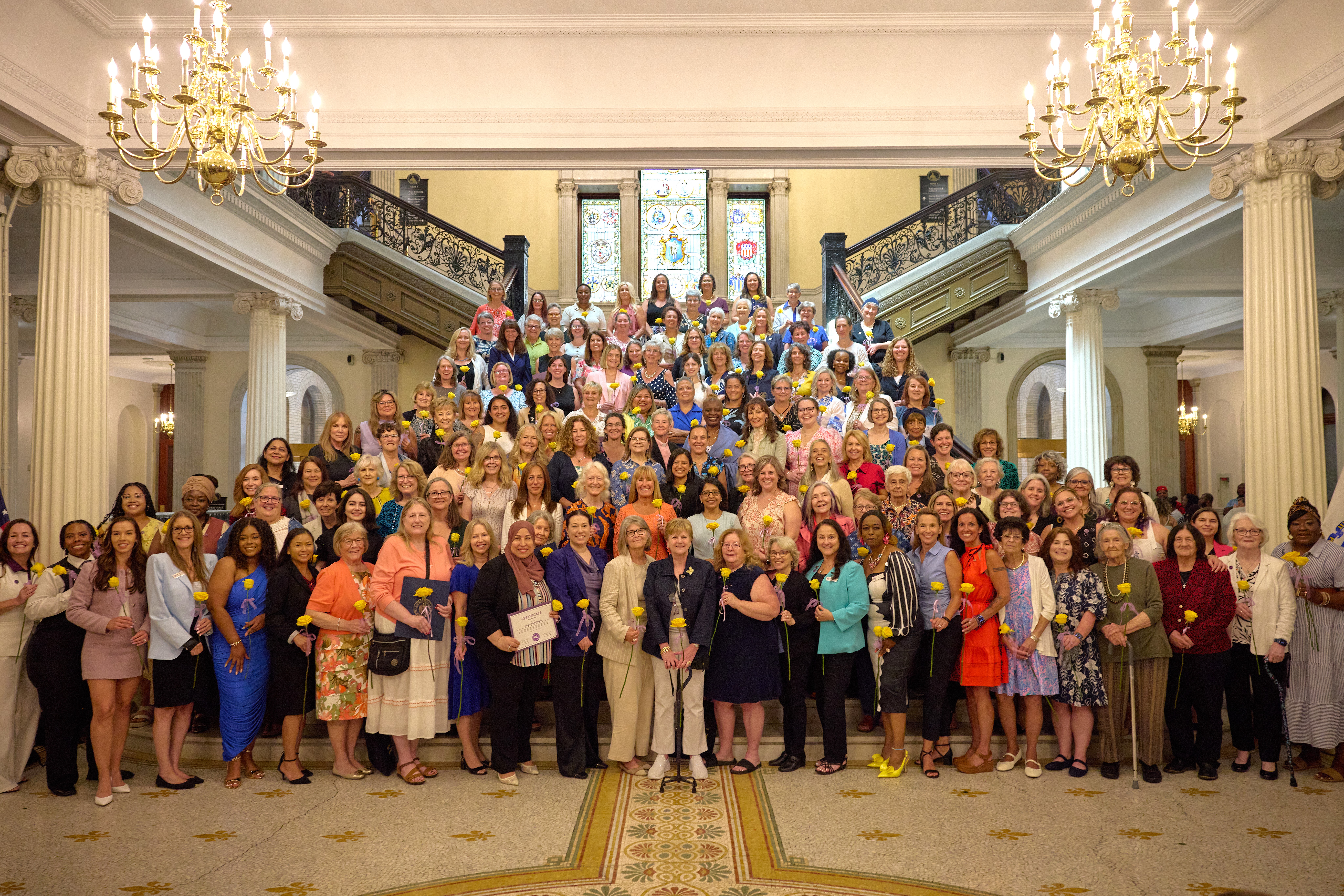 A large and diverse group photo of more than 147 women, the Class of 2026 Commonwealth Heroines, on the steps of the Grand Staircase at the Massachusetts Statehouse.